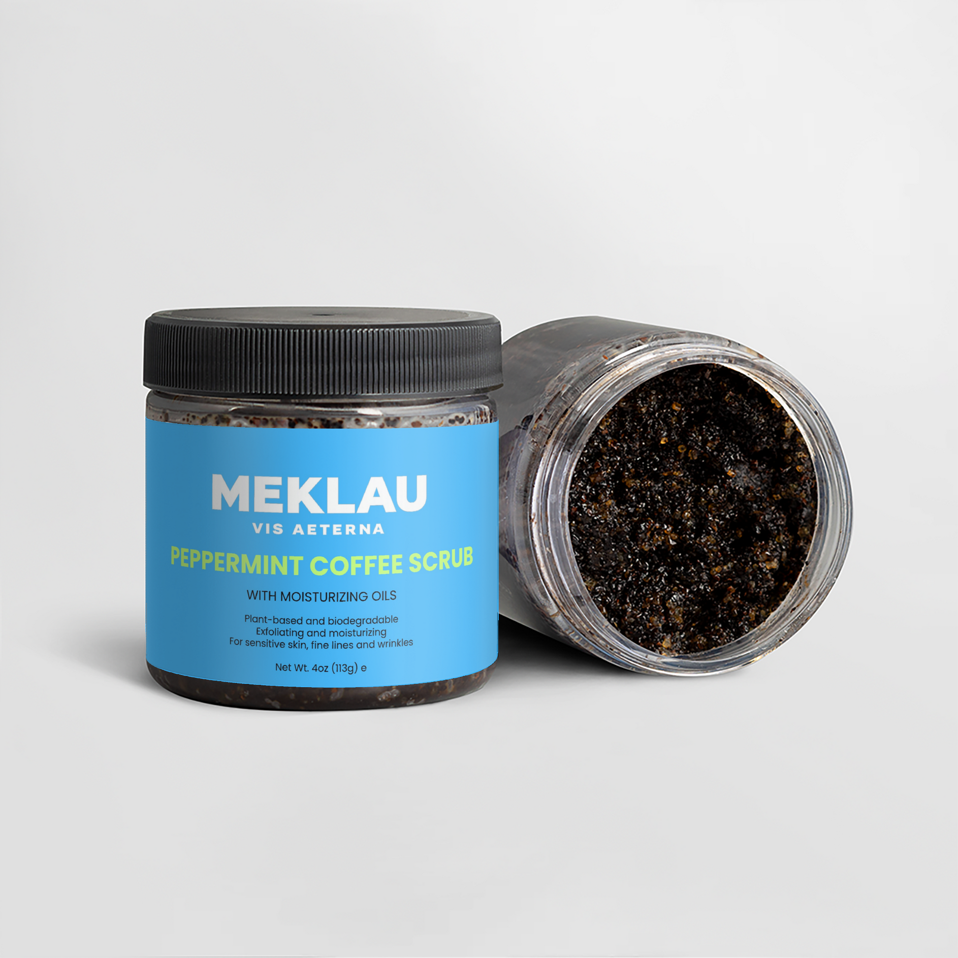 Two jars of Meklau Vis Aeterna Peppermint Coffee Scrub on a light gray background, with one jar standing upright showing the blue label and the other lying down to show the dark coffee scrub inside.