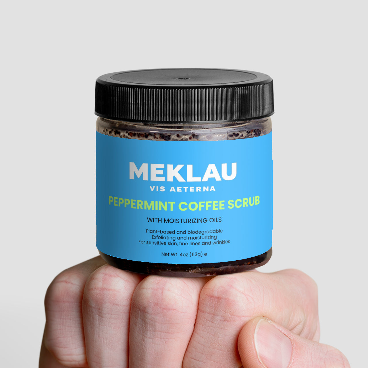 A hand holding a jar of MEKLAU Vid Asterna Peppermint Coffee Scrub against a light gray background.