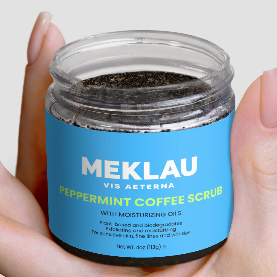 A hand holding a clear jar of MEKLAU Vis Aeterna Peppermint Coffee Scrub, showing the dark coffee grounds inside and a bright blue label with white and yellow text.